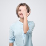 European young woman in blue shirt closing one eye with hand and smiling.